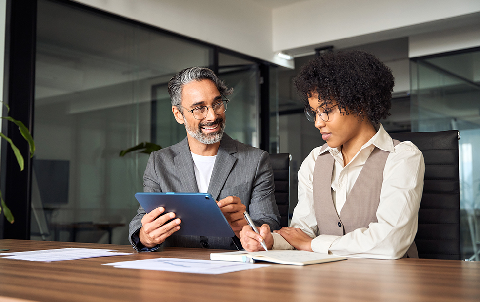 Alt text: Two professionals reviewing a tablet and taking notes together at a conference table.